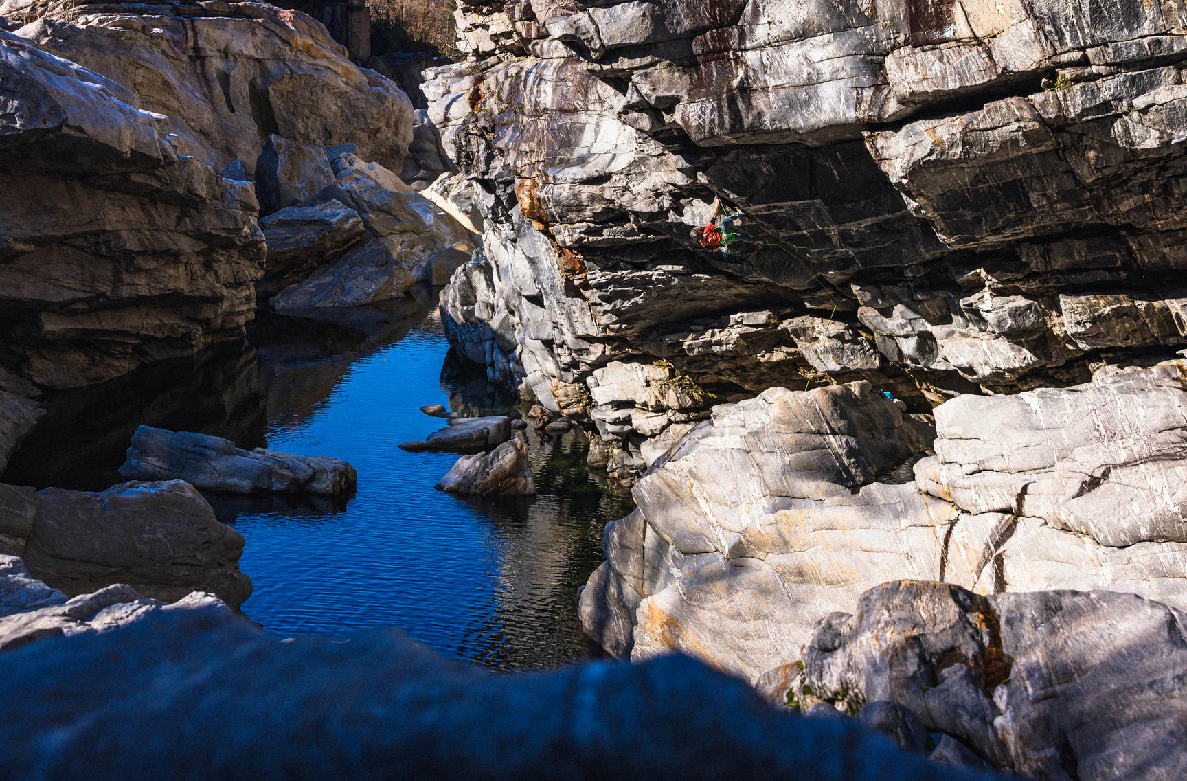 man climbing a rocky cliff wearing e9 clothing with a body of water below